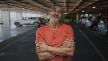 Fototapeta premium Man with arms crossed, hands visible, standing in an indoor parking building beside parked cars; confidence resilience.