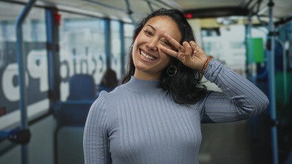 Woman makes peace sign with hand at eye in street bus interior, smiling and showing hoop earring  joy carefree energy. © Krakenimages.com