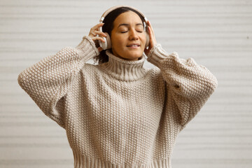 A serene woman with closed eyes wearing a beige turtleneck knitted sweater and wireless headphones,...