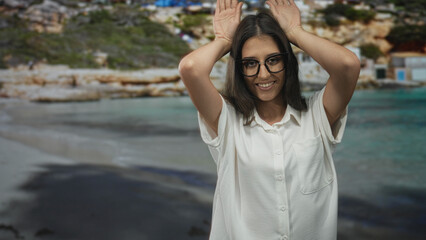Woman with hands raised to head making antlers gesture on street beside beach and sea, wearing...