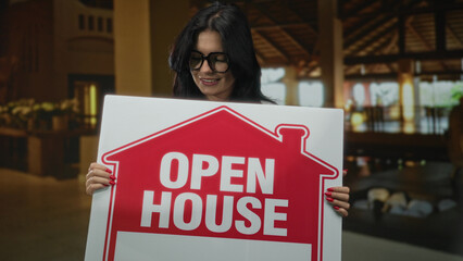 Woman with glasses stands smiling and holds red open house sign in hotel building  home sale joy marketing. © Krakenimages.com