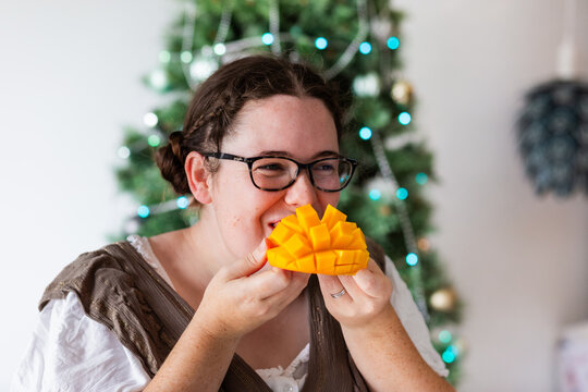 Adult woman standing indoors by Christmas tree eating fresh mango fruit
