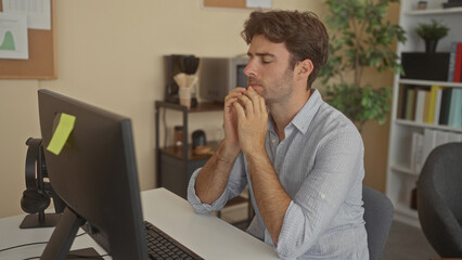 Young hispanic man at computer monitor with hands on temples in office building; stress...