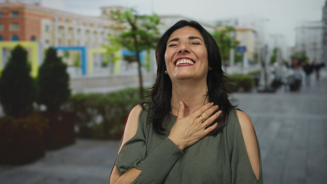 Middle age hispanic woman laughing with hand on cheek on sunlit bustling street; warmth joy positivity.