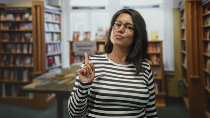 Woman touching her glasses and pointing a finger toward the viewer amid bookshelves in a library building, wearing a striped top  thoughtful. © Krakenimages.com
