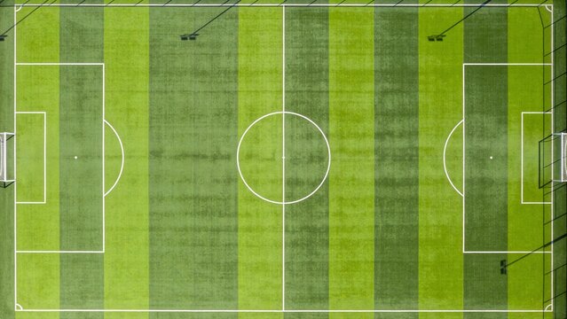 Image is a top-down view of a standard soccer field, featuring a symmetrical layout with clearly marked white lines on a vibrant green grass surface. The field is divided into two halves by a central 