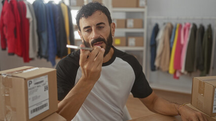 Fototapeta premium Man holds phone to mouth while packing cardboard box for shipping amid clothing racks and shelves in studio; focused entrepreneurship.