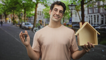 Fototapeta premium Man wearing glasses and a pale pink shirt holds a keyring and wooden house model, smiling and holding keys forward on a tree-lined city street with parked cars; homeownership confidence.