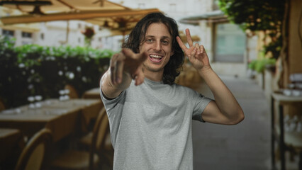 Fototapeta premium Man young hispanic pointing and flashing peace sign with hand on street cafe terrace outdoors; playful energy.
