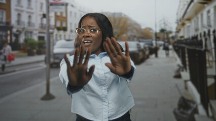 Fototapeta premium Black woman wearing glasses raises hands in a defensive gesture on a city street, facing oncoming traffic; apprehension caution.
