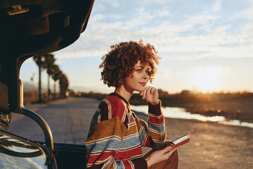 Young woman with curly hair wearing rainbow sweater stands near car at sunset holding smartphone and book looking thoughtfully into distance outdoors on empty road. © SHOTPRIME STUDIO