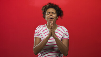 Young african american woman pressing hands together in prayer in studio against red backdrop; hope...