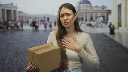 Obraz premium Woman holding cardboard box, hand on chest, anxious facial expression and long hair, casual top in crowded street; worry delivery problem.