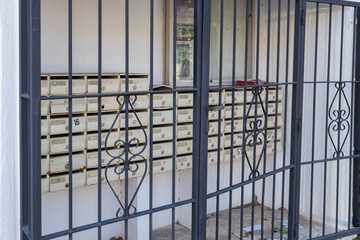 A weathered dark grey metal gate, featuring elegant decorative scrollwork, stands partly open, revealing a collection of aged, cream-colored wall-mounted mailboxes