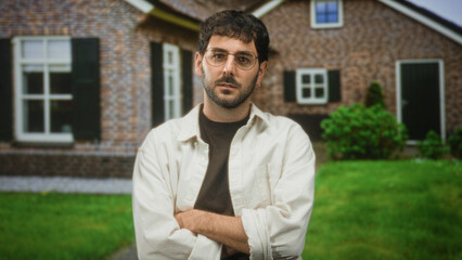 Hispanic man with beard and glasses stands with arms crossed, looking aside on a green lawn in front of a brick residential building wearing a casual shirt  pensive quiet reflection. © Krakenimages.com