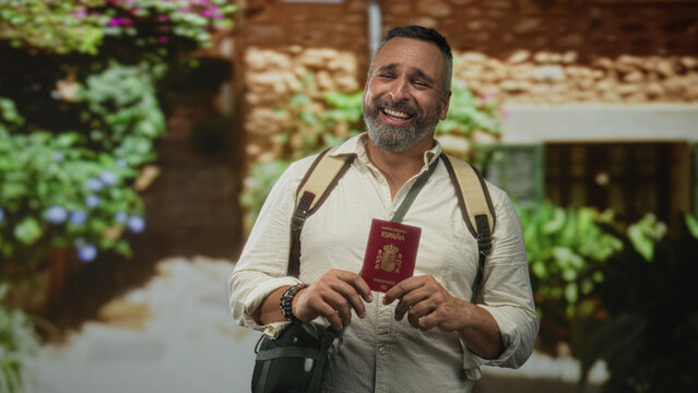 Man holding passport and smiling with hand raised beside backpack at building entrance of a sunlit building outdoors; travel joy.