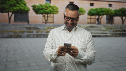 Fototapeta premium Man with beard smiling while tapping smartphone with hands on old town street; digital connection contentment.