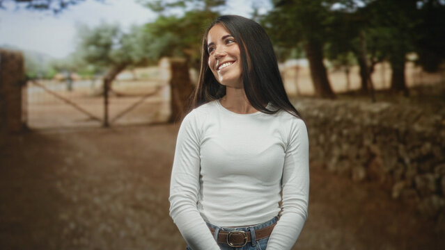Woman smiling, hands clasped, looking up beside a rustic gate on a street in white long sleeve top and jeans; serene contentment.