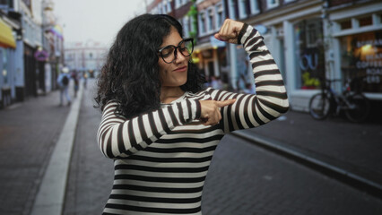 Fototapeta premium Woman flexes arms and points to bicep on a bustling city street with shopfronts and bicycle in frame; confidence empowerment pride.