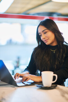 Young Latina woman working on laptop in modern cafe with coffee cup, focused female typing on computer while enjoying relaxed workspace and warm indoor atmosphere