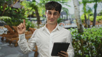 Fototapeta premium Man points finger to tablet while holding it in building near lounge chairs and palm trees, smiling; friendly guidance.