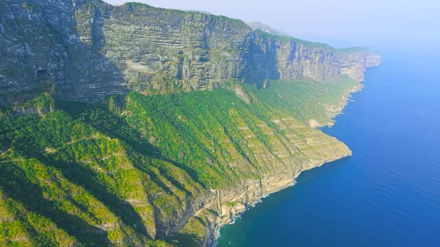 Aerial view of steep green mountains and vertical rock cliffs meeting the blue ocean waters at Jabal Shaat, Oman and Yemen border.