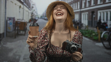 Fototapeta premium Redhead woman holding coffee cup and vintage camera, laughing with open mouth on cobblestone street, straw hat and floral dress visible; joyful travel moment.