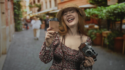 Redhead woman laughing, holding a takeaway coffee cup and vintage camera, wearing straw hat and...