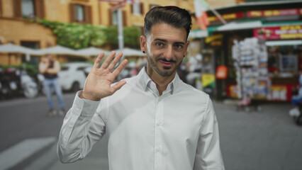 Obraz premium Young hispanic man wearing white shirt waving hand on busy urban street sidewalk in bright daylight; friendliness.