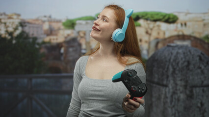 Redheaded woman holding a gamepad and wearing blue headphones, head tilted and smiling in front of a building  playful. © Krakenimages.com