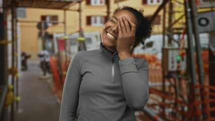 Fototapeta premium Young african american woman laughing and covering her face with hand at an outdoor building construction site scaffolding; joy.