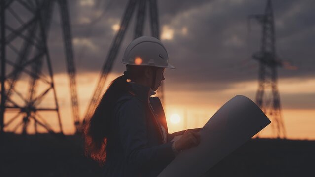 An electrician on the background of high towers of power plants looks at the project for the development of an electrical structure, the expansion of the electrical voltage of volts in the wires.