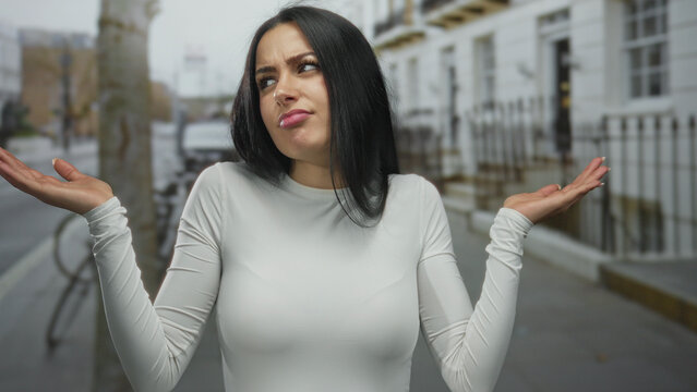Woman on city street with a puzzled expression, wearing a white shirt, gestures questioningly against an urban backdrop, expressing confusion on a busy street outdoors.