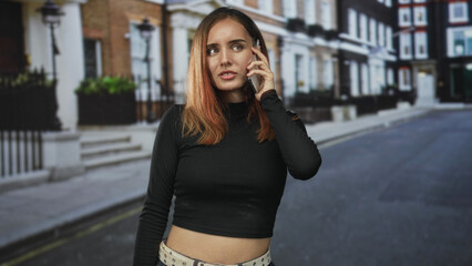 Fototapeta premium Redhead woman holding smartphone to ear on street, black crop top and studded belt visible while she looks worried and glances aside; concern urgency.