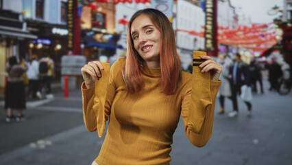 Fototapeta premium Young redhead woman holds mustard socks with raised arms on a busy city street, smiling and showing hosiery to camera; cozy playful.
