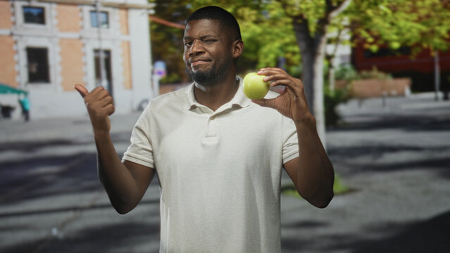 Young black man holding green apple with hand and thumb pointing left on a street; skepticism choice.