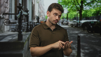 Fototapeta premium Young hispanic man wearing a brown polo pointing a finger to his palm on a leafy city street, smiling while tapping his open hand; casual happiness.