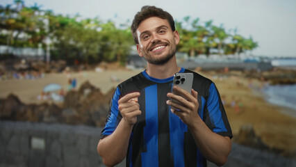 Young hispanic man points finger to smartphone on beach while smiling and holding phone in hands;...