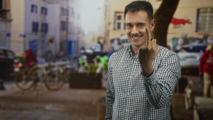 Fototapeta premium Hispanic man with moustache shows middle finger flipping gesture in street wearing plaid shirt and smiling; defiance cheeky.