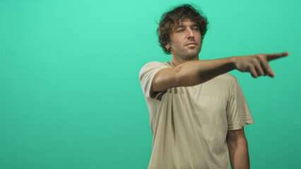 Fototapeta premium Man points finger forward with outstretched arm and visible hand, wearing a beige t shirt against green studio backdrop; determination.