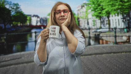 Fototapeta premium Woman holds mug reading i am the boss while touching her forehead on a street canal bridge scene; confidence humor.