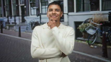 Fototapeta premium Man with hand on chin and slight smile, wearing cream sweater, standing on cobblestone street by building; thoughtful calm confidence.