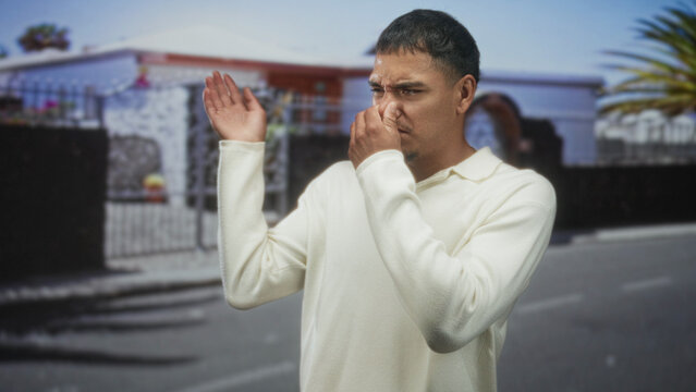 Man pinches nose in white sweater on a residential street while reacting to a strong odor; disgust odor.