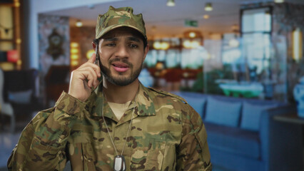 Fototapeta premium Young soldier in camouflage uniform holds smartphone to ear standing by front desk in hotel lobby; concern duty.