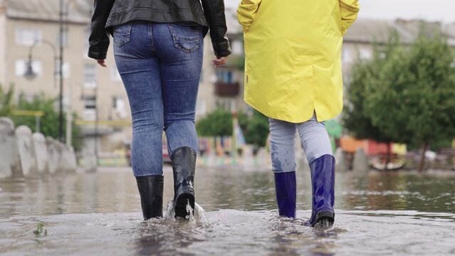 girls in rubber boots are walking in a puddle, a flood on a city street, flood on the sides of roads and asphalt roadway, splashes different directions from wet muddy puddle, presence water districts.