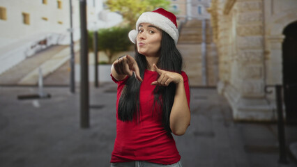 Fototapeta premium Young woman wearing santa hat points finger to camera on street, smiling and posing with visible hands; holiday cheer playful.