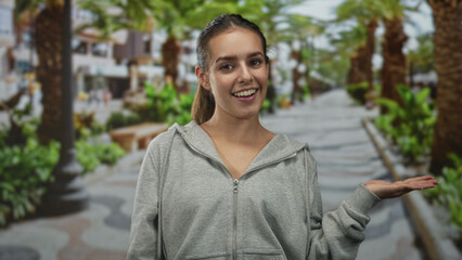 Obraz premium Woman smiling and holding palm up in gray hoodie on street promenade showing open hand gesture; happy friendly welcome.