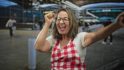 Fototapeta premium Senior hispanic woman cook in red gingham apron raises clenched fists and laughs with eyes closed in airport terminal; joy celebration victory.