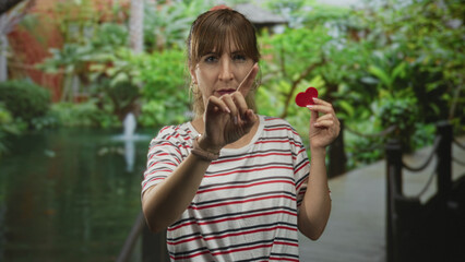 Fototapeta premium Woman points finger while holding a small red heart near chest on park walkway by green pond and bridge; love longing.
