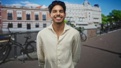 Fototapeta premium Man smiling and clasping hands beside parked bicycles on a street with historic buildings and a canal, facing camera and leaning slightly forward; joy gratitude welcome.